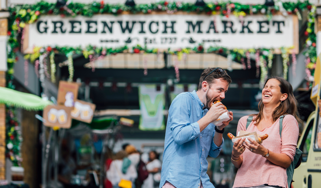 A man and woman laugh while eating sausages at Greenwich Market